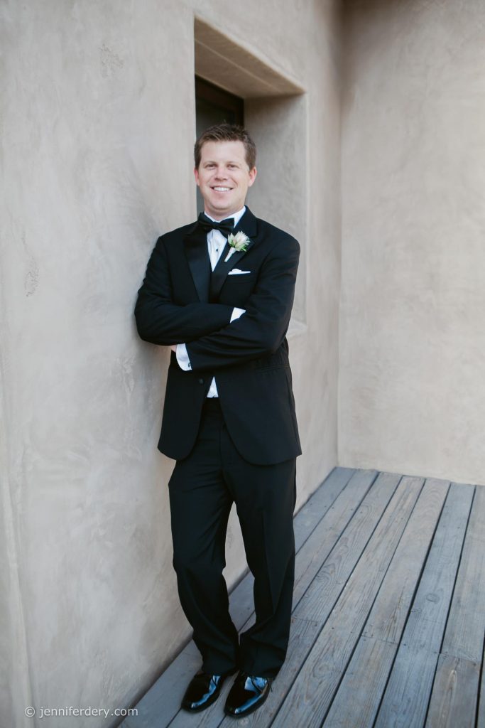 A man in a black tuxedo with a bow tie and white boutonniere stands smiling with arms crossed, leaning against a beige stucco wall on a wooden deck.
