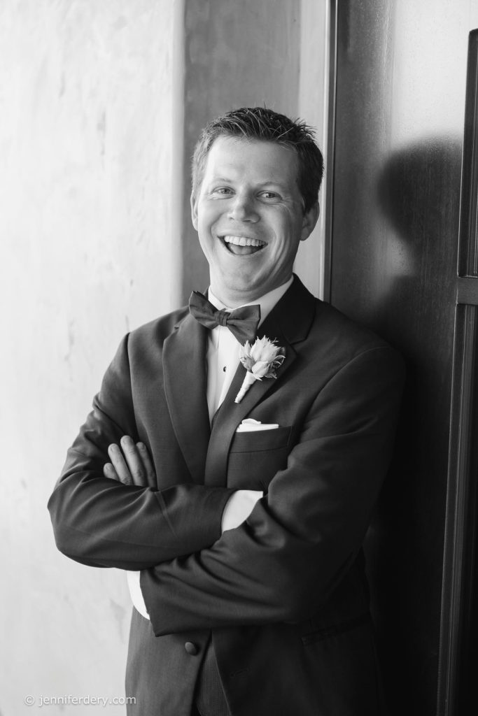 A man in a tuxedo with a bow tie and boutonnière stands with arms crossed, smiling and leaning against a wall. The photo is in black and white.