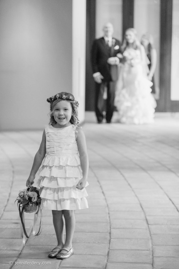 A young girl in a ruffled dress and flower crown stands smiling with a basket of flowers. In the blurred background, two adults and a bride in a wedding gown walk toward her.
