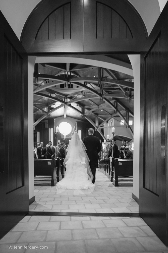 A bride in a flowing gown walks down the aisle with a man, likely her father, in a church filled with guests. The scene is viewed from behind through large open doors.