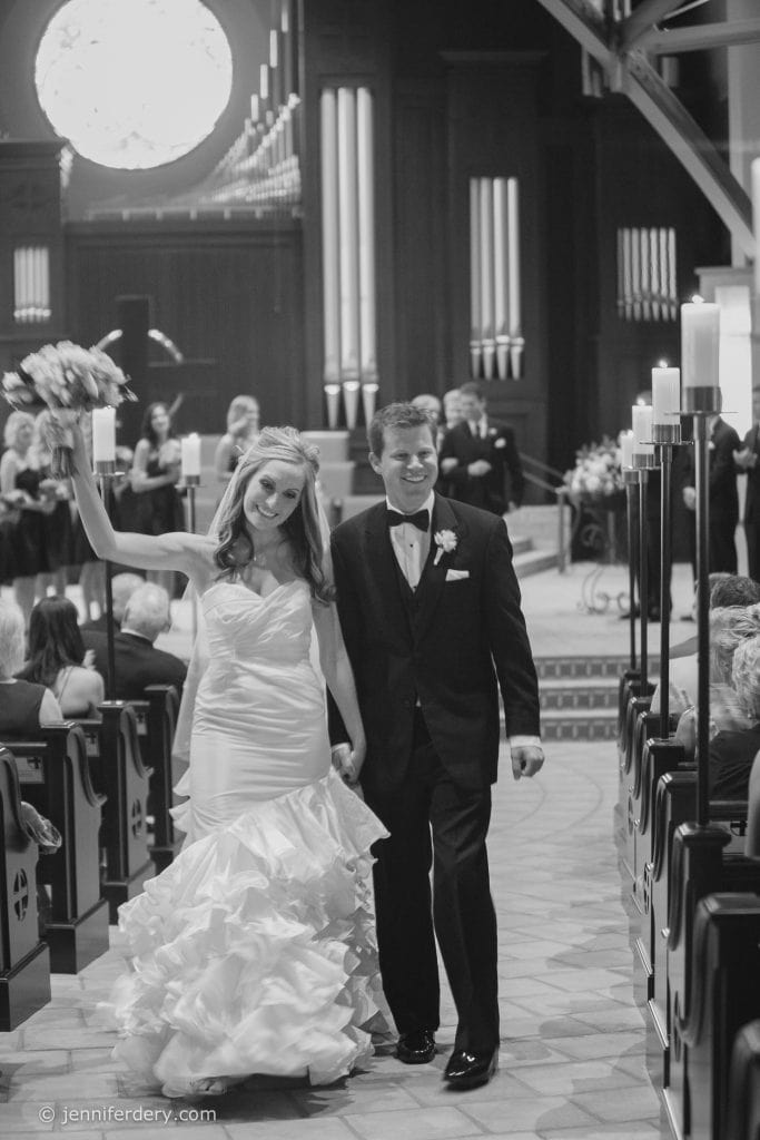 A bride and groom walk down the aisle of a church, smiling happily. The bride holds a bouquet aloft, and guests sit in pews on either side. The scene is elegant and joyful, captured in black and white.