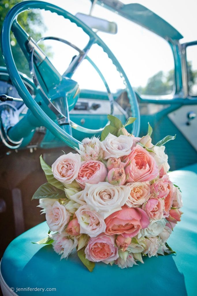 A bouquet of pink and white roses rests on the turquoise seat of a vintage car, with sunlight streaming in through the window and the steering wheel visible in the background.