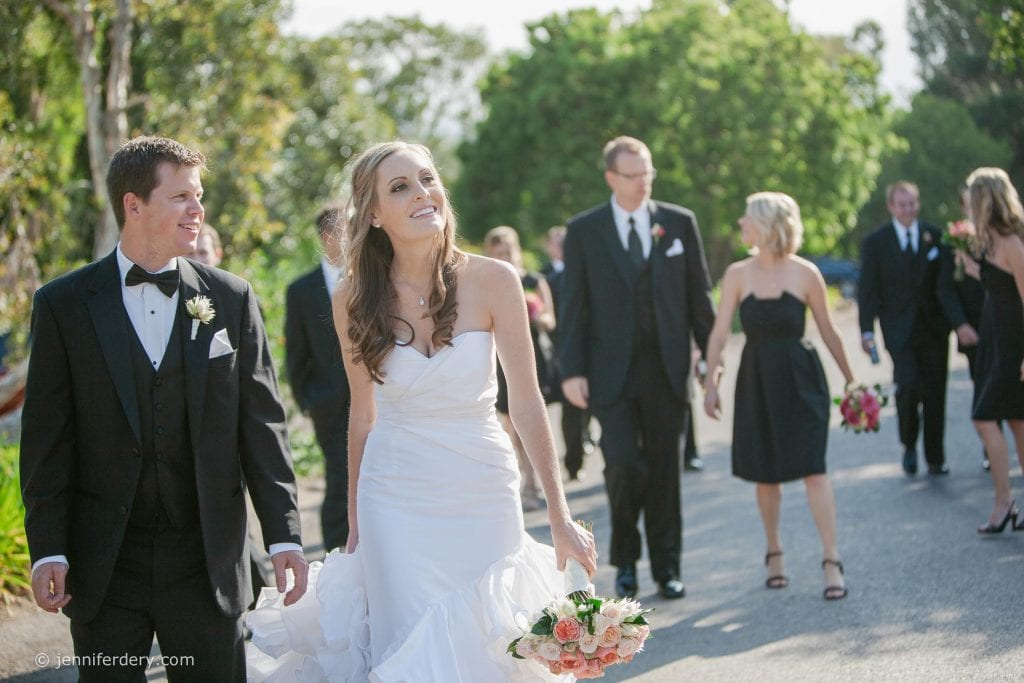 A smiling bride in a white dress walks beside the groom in a black tuxedo. They are outdoors with bridesmaids and groomsmen in black attire walking behind them. The bride holds a bouquet of flowers.