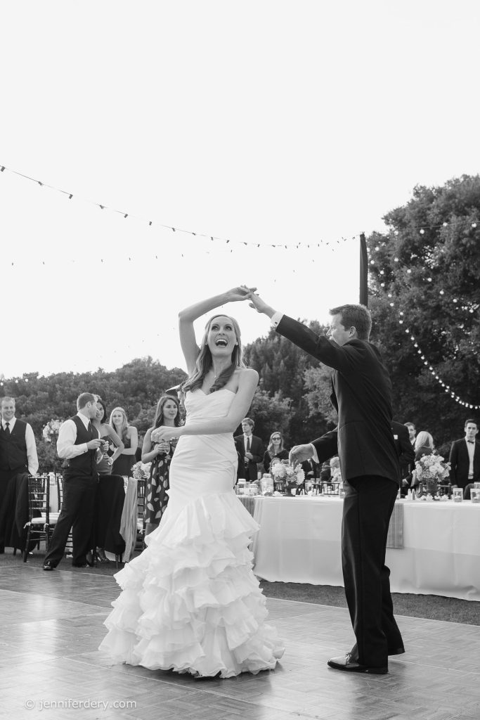 A bride and groom dance outdoors on a wooden floor, smiling and surrounded by guests. The bride wears a ruffled dress; string lights and trees are in the background. The photo is in black and white.