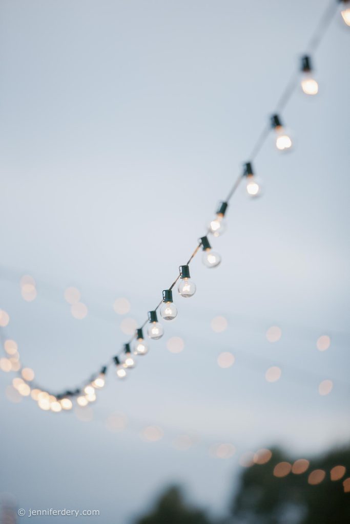A string of round, white outdoor lights hangs diagonally against a soft, light blue sky, with more blurred lights in the background creating a dreamy, festive atmosphere.