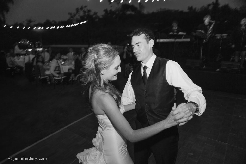 A bride and groom dance together outdoors at night, surrounded by guests seated at tables, with string lights overhead and a live band playing in the background.