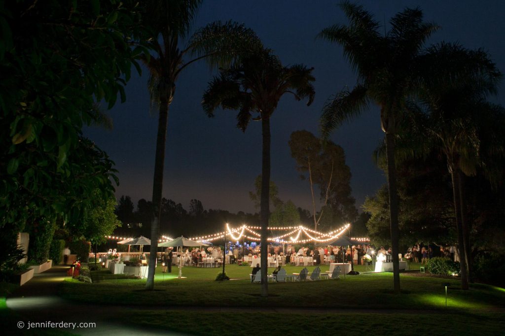 An outdoor evening event with tables and chairs set up on the croquet lawn at wedding at Rancho Valencia Resort, illuminated by string lights draped between palm trees. Guests are gathered under the lights, surrounded by trees and greenery.