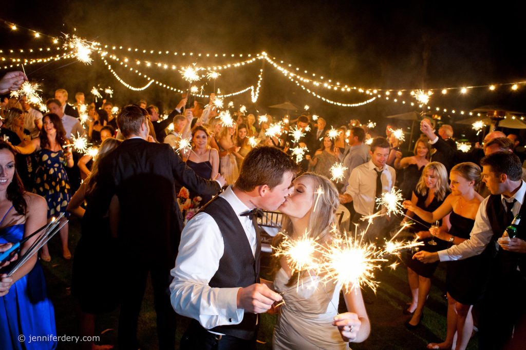 A couple kisses while holding sparklers at an outdoor night wedding at Rancho Valencia Resort, surrounded by guests also holding sparklers. String lights hang above, creating a festive atmosphere.