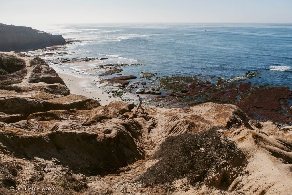 A person walks along rugged, sandy cliffs overlooking a rocky shoreline and the blue ocean under a clear sky. Coastal vegetation and tide pools are visible below.