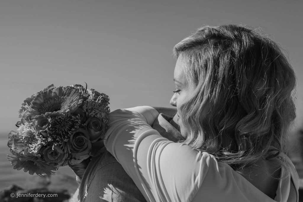 A woman with wavy hair hugs someone while holding a bouquet of flowers. The image is in black and white and appears to be taken outdoors, possibly at a wedding or special event.