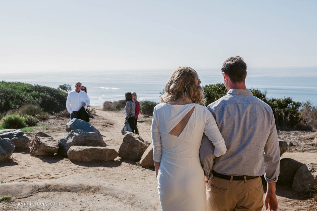 A couple, seen from behind, walks arm in arm toward a scenic coastal overlook, where three people stand near large rocks. The ocean and clear sky are in the background.