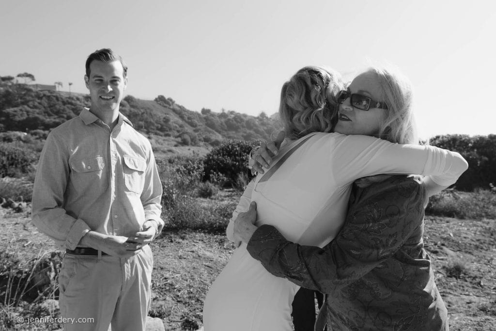 A man smiles at the camera while two women embrace in a hug outdoors, with hills and shrubs in the background. The scene is captured in black and white.