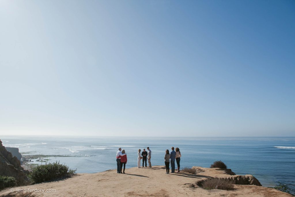 A group of people stands on a sandy cliff overlooking the ocean under a clear blue sky, with waves and coastline visible in the distance.