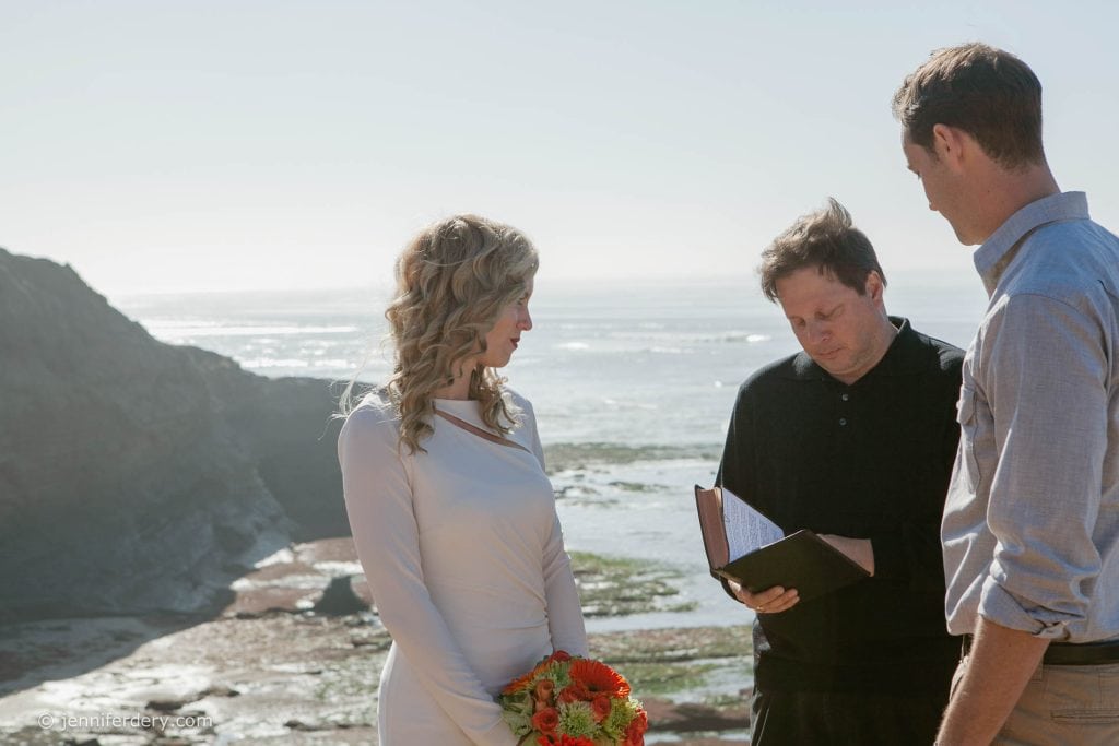 A bride in a white dress holding a bouquet stands beside a groom as an officiant reads from a book during a sunny beachside wedding ceremony, with cliffs and ocean in the background.