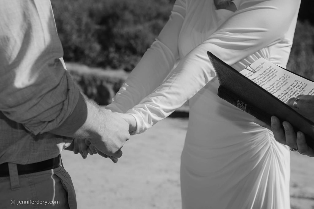 A bride and groom hold hands during an outdoor wedding ceremony as an officiant reads from a book. Only their torsos and hands are visible in the black-and-white photo.