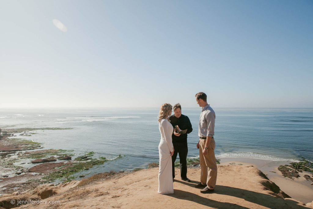 A couple stands on a sandy cliff overlooking the ocean, facing an officiant who is holding a book, as they appear to be getting married under clear blue skies.