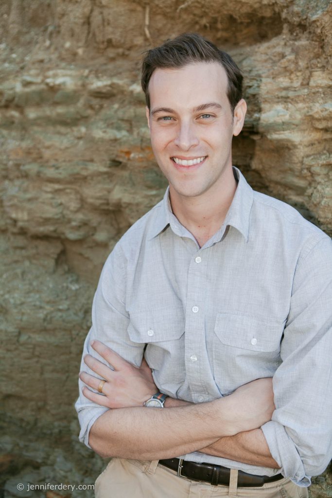 A man with short brown hair, wearing a light gray button-up shirt, stands in front of a rocky wall, smiling with his arms crossed.