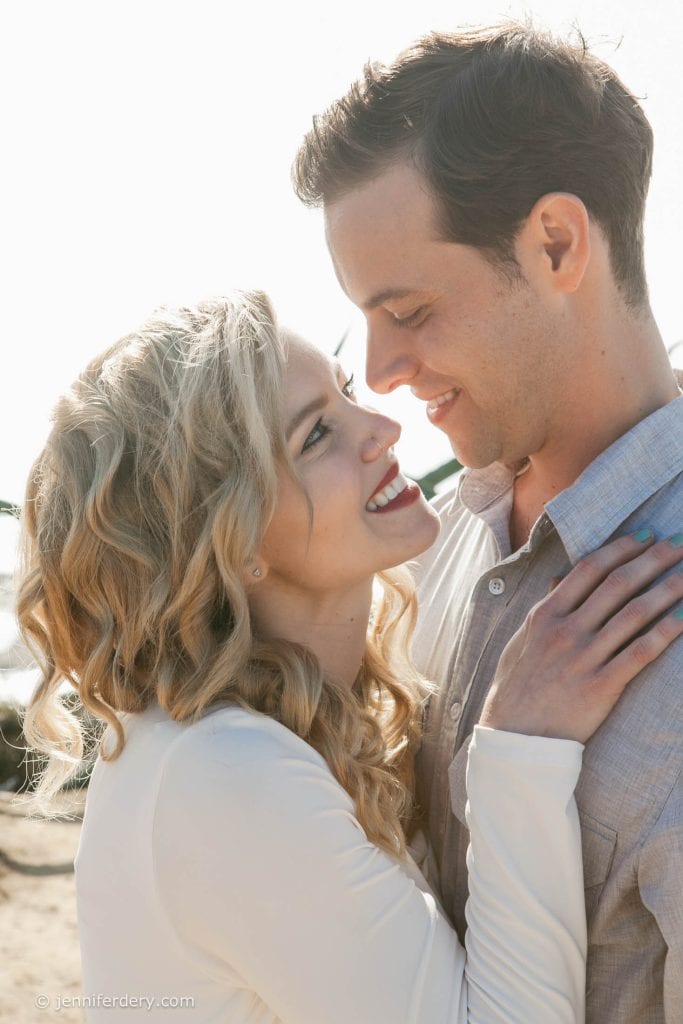 A couple stands close together outdoors, smiling and gazing into each other's eyes. The woman with wavy blonde hair and red lipstick holds the man's shoulder, while he smiles warmly at her under bright sunlight.