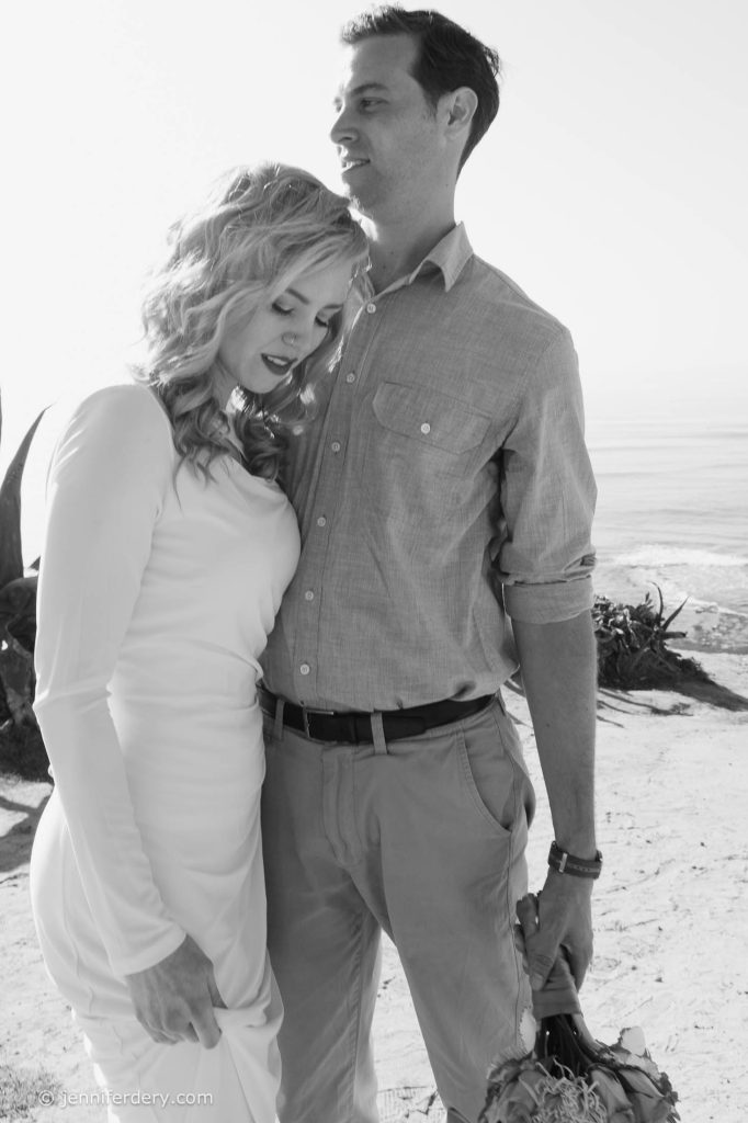 A woman in a white dress rests her head on a man's chest as he gently holds her, both standing on a beach. The scene is in black and white, with the ocean and bright sky in the background.