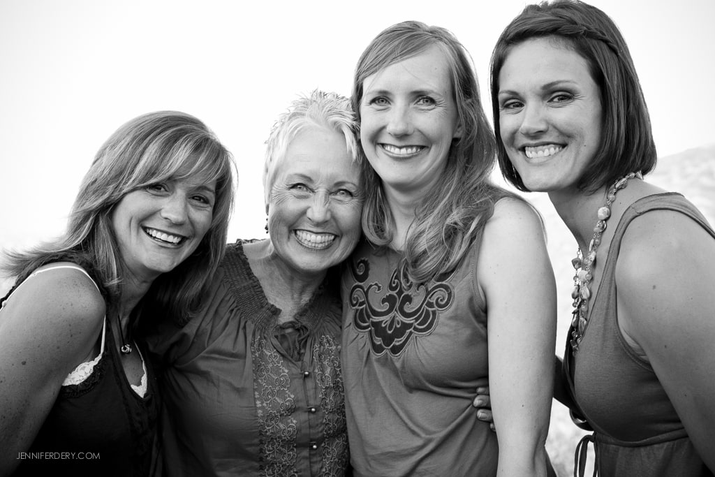 Four women smiling and standing close together in a black and white photo. They are outdoors with a natural, blurred background.