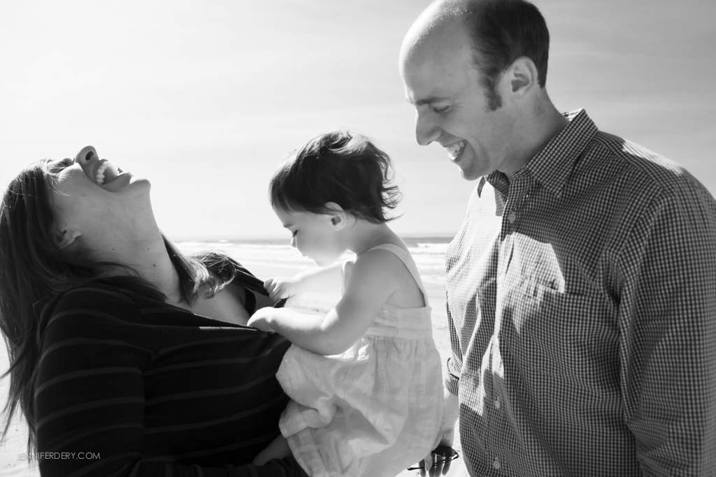 A joyful family moment at the beach in black and white. A mother is laughing while holding a toddler, and the father stands beside them, smiling. The background shows a clear sky and the ocean.