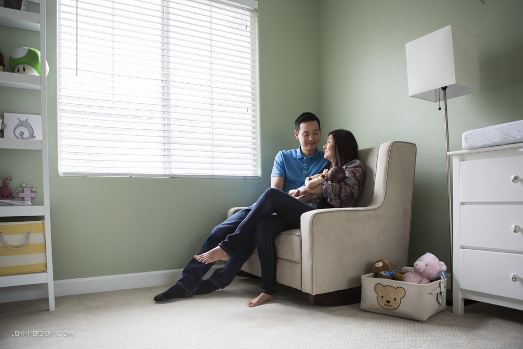 A couple sits together in a cozy nursery room, holding a baby. The room is painted green with white blinds, featuring a bookshelf, a changing table, and a basket with stuffed animals near a white dresser. Soft lighting adds a warm atmosphere.