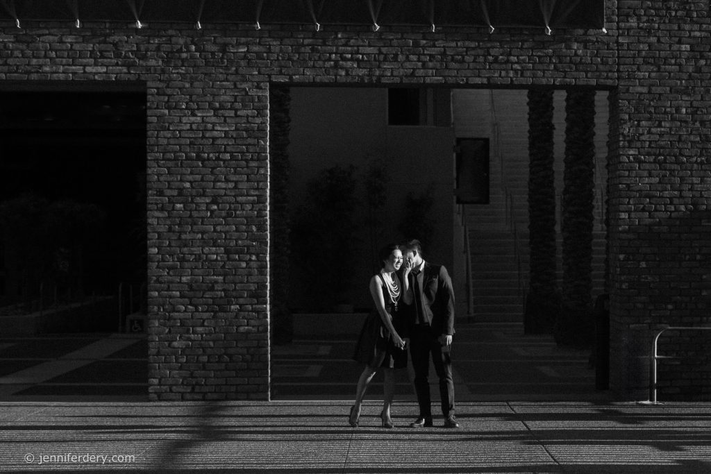 A couple dressed formally stands close together in the shadow of a large brick structure, with sunlight highlighting their faces and casting long shadows on the ground.
