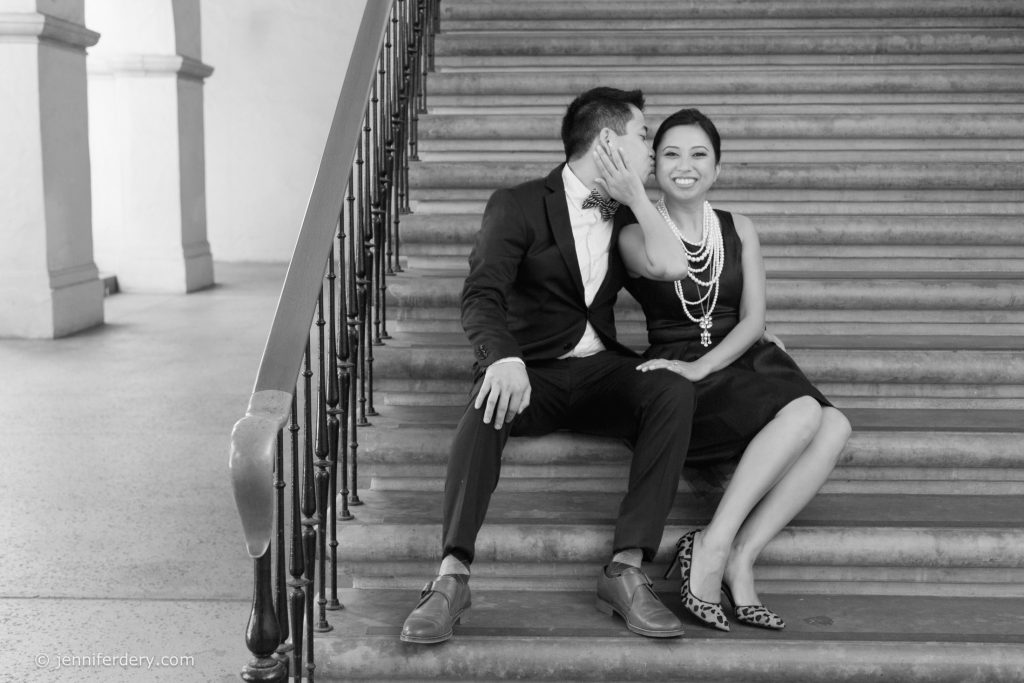 A man in a suit and a woman in a dress sit on stairs. The man kisses the woman's cheek while she smiles brightly at the camera, her hand touching his face. They appear joyful and affectionate.