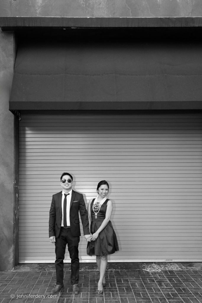 A man in a suit and sunglasses stands holding hands with a woman in a dress and necklace, both smiling, in front of a closed metal shutter and under a large black awning. The scene is in black and white.