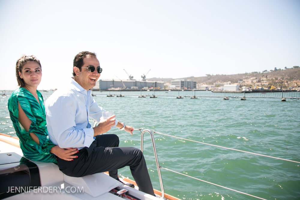 A woman in a green dress sits on a boat next to a smiling man in a light blue shirt and sunglasses, with docks and industrial buildings visible across the water in the background.