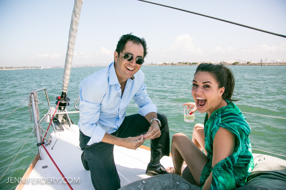 A man in sunglasses and a light blue shirt and a woman in a green dress smile and laugh while relaxing on a sailboat, with water and a distant shoreline in the background.