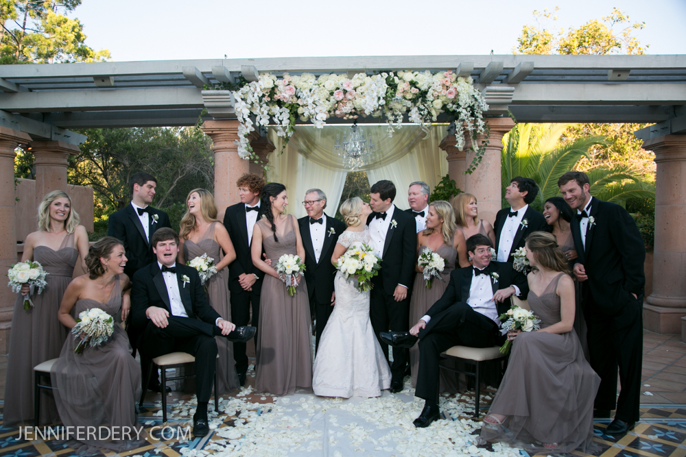 A wedding party poses outdoors under a flower-adorned pergola. The bride and groom stand in the center, surrounded by bridesmaids in taupe dresses and groomsmen in black suits, all smiling and interacting joyfully.