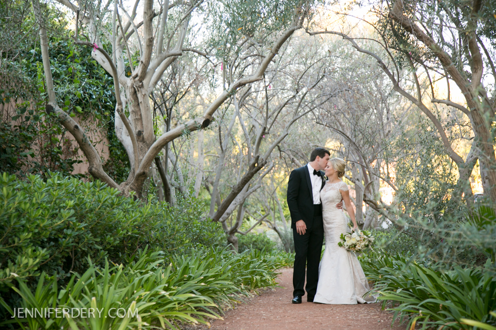 A bride and groom share a kiss while walking down a tree-lined garden path, surrounded by lush greenery. The bride holds a bouquet, and the groom wears a tuxedo. Sunlight filters through the trees.