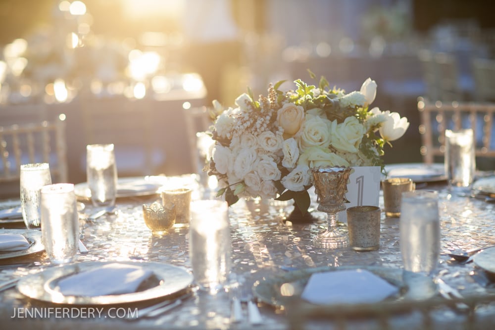 A sunlit outdoor table set for an event features a floral centerpiece with white roses, gold-rimmed glassware, and plates, with shimmering sunlight reflecting off the glasses and table.