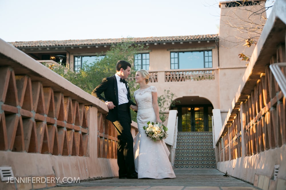 A bride and groom stand close together on a sunlit bridge outside a Mediterranean-style building. The bride holds a bouquet, and both are smiling, dressed in formal wedding attire.