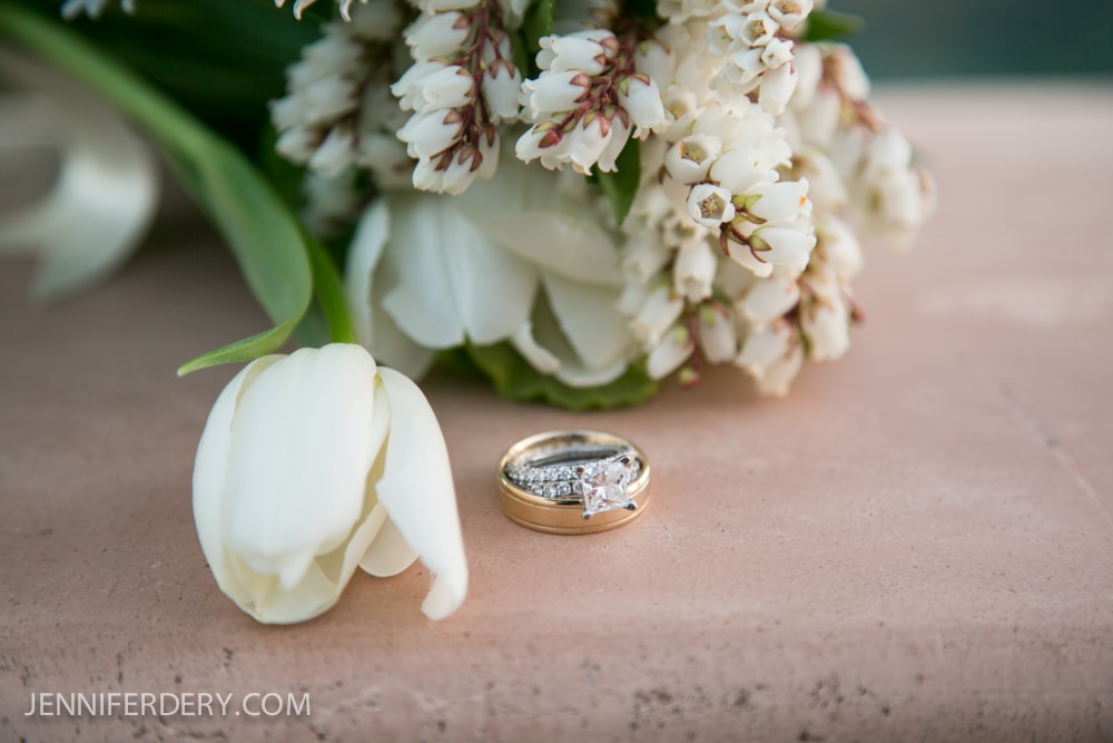 A close-up of a white tulip, a small bouquet of white flowers, and a pair of wedding rings with a diamond engagement ring resting on a stone surface. The photo credit "JENNIFERDERY.COM" appears in the bottom left corner.