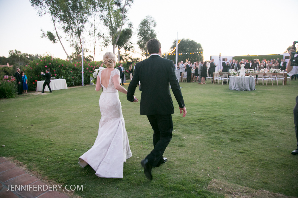 A bride and groom walk hand in hand across a grassy lawn at Rancho Valencia Resort toward a crowd of wedding guests at an outdoor reception, with trees and string lights in the background.