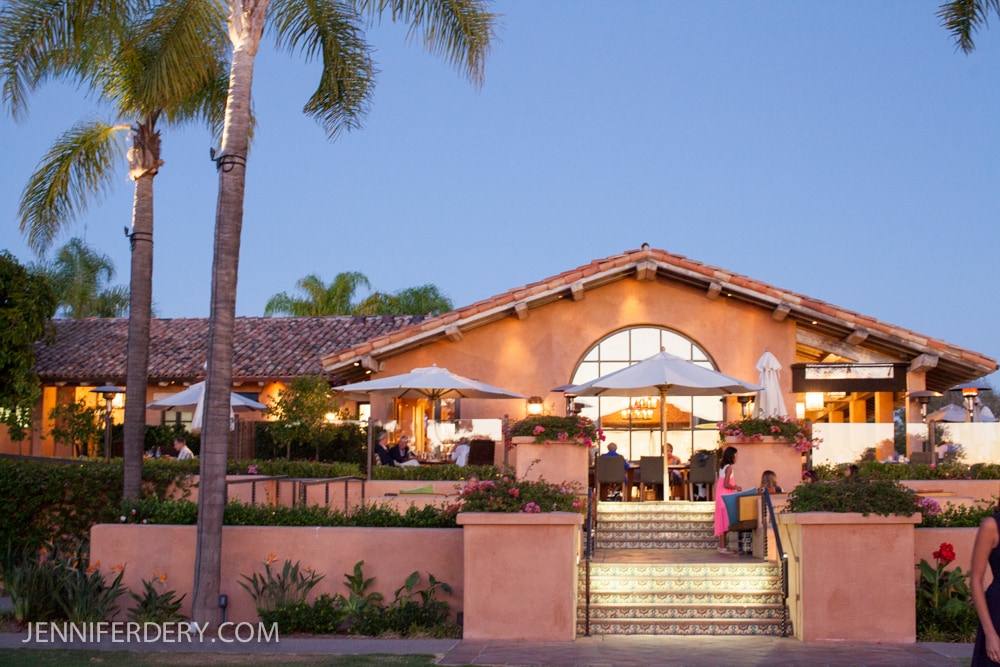 Rancho Valencia Resort: A Spanish-style villa with a terracotta roof and arched windows, surrounded by palm trees and vibrant gardens. Patio dining tables with umbrellas are visible, and tiled steps lead to the entrance at dusk.