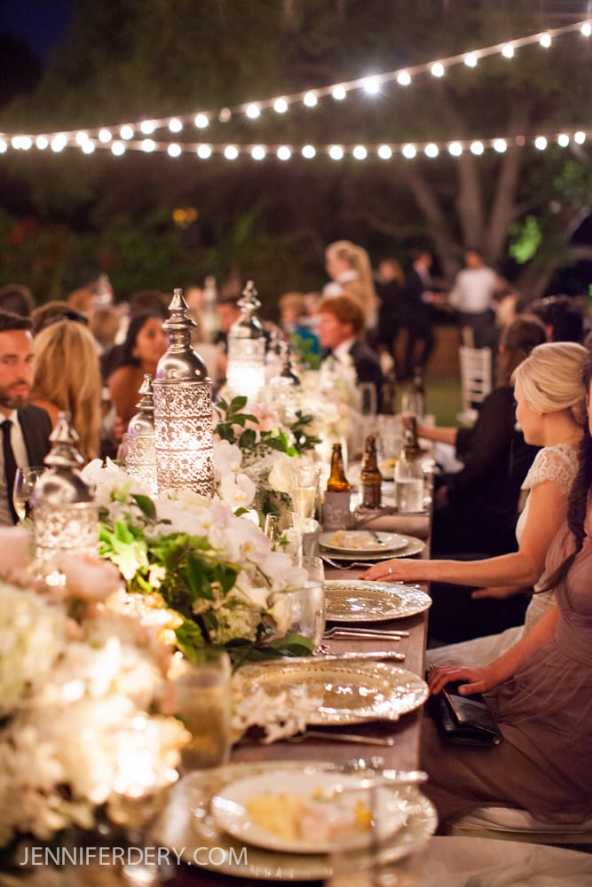 An elegant outdoor dinner party at night at Rancho Valencia Resort with guests seated at a long table decorated with flowers, candles, and lanterns under string lights. The atmosphere is festive and warm.