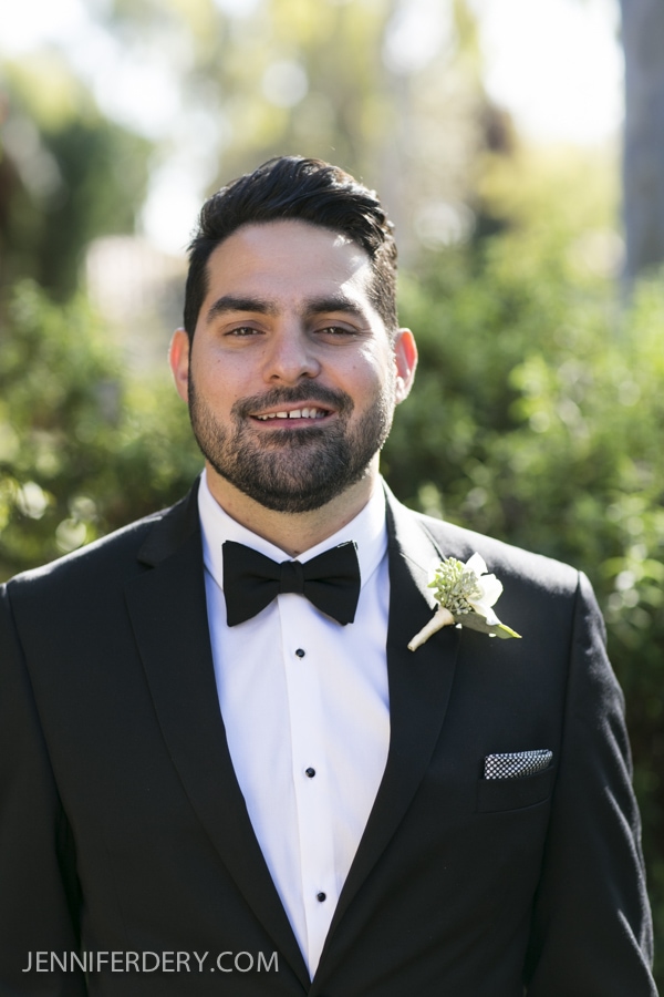 A man in a black tuxedo with a black bow tie and white boutonniere smiles outdoors, standing in front of greenery at an Estancia La Jolla Wedding. The photo is well-lit and formal.