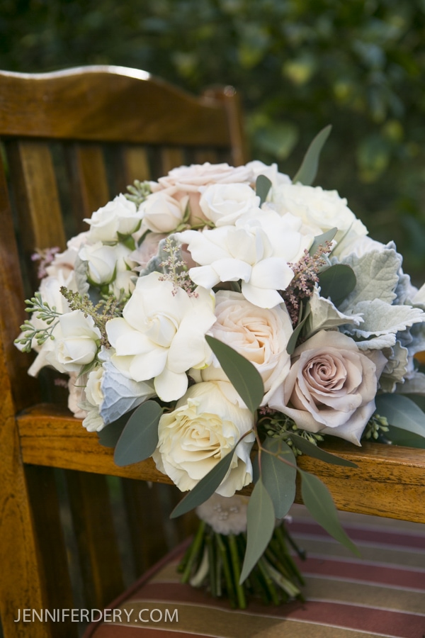 A bouquet of white and pale pink roses with greenery sits on a wooden chair with a striped cushion, capturing the elegance of an Estancia La Jolla Wedding. The background is blurred foliage.