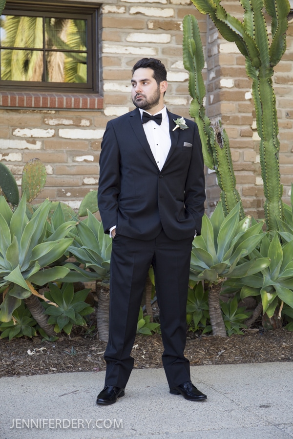 A man in a black tuxedo stands outside in front of a brick wall and lush greenery, including cacti and succulents, at an Estancia La Jolla Wedding. He gazes to the side with his hands tucked in his pockets.