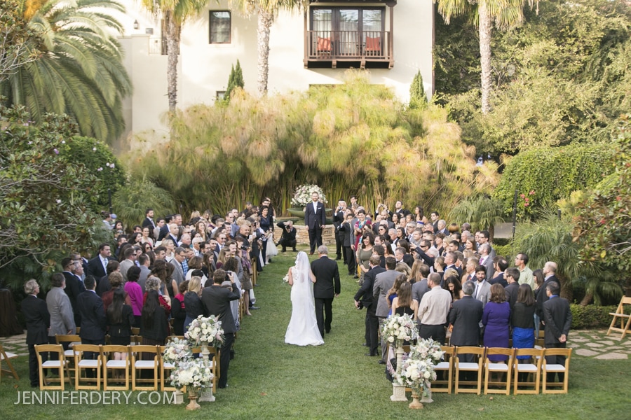 An outdoor Estancia La Jolla Wedding ceremony with a bride and her escort walking down the aisle toward the groom. Guests are seated on both sides, surrounded by lush greenery, flowers, and palm trees on a sunny day.