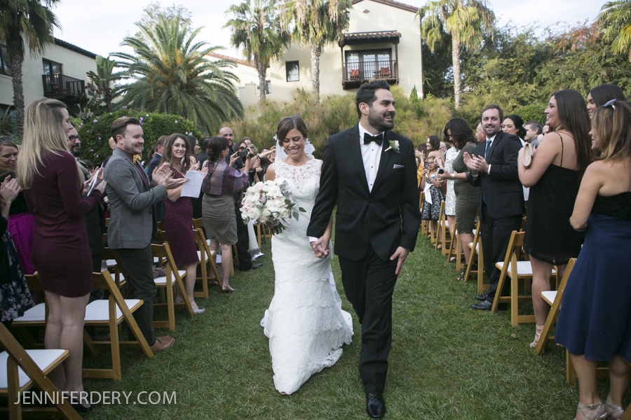 A bride in a white dress and groom in a black tuxedo walk down an outdoor aisle at their Estancia La Jolla Wedding, smiling and holding hands, as wedding guests seated on either side stand and applaud.