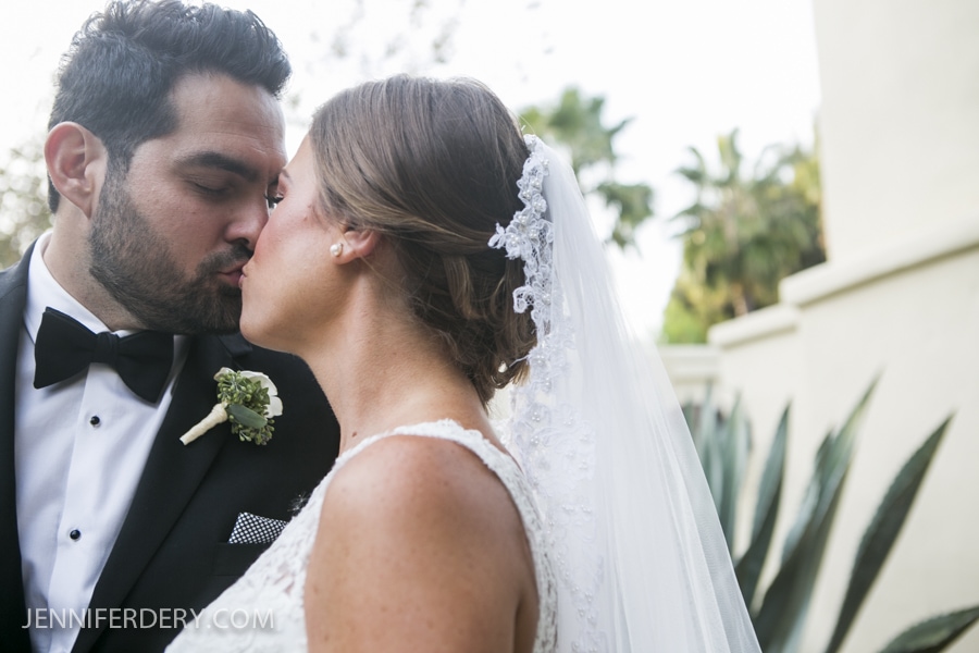 A bride and groom share a kiss outdoors on their Estancia La Jolla Wedding day. The groom wears a black tuxedo, and the bride wears a white lace dress with a veil. Greenery and a light-colored wall are in the background.