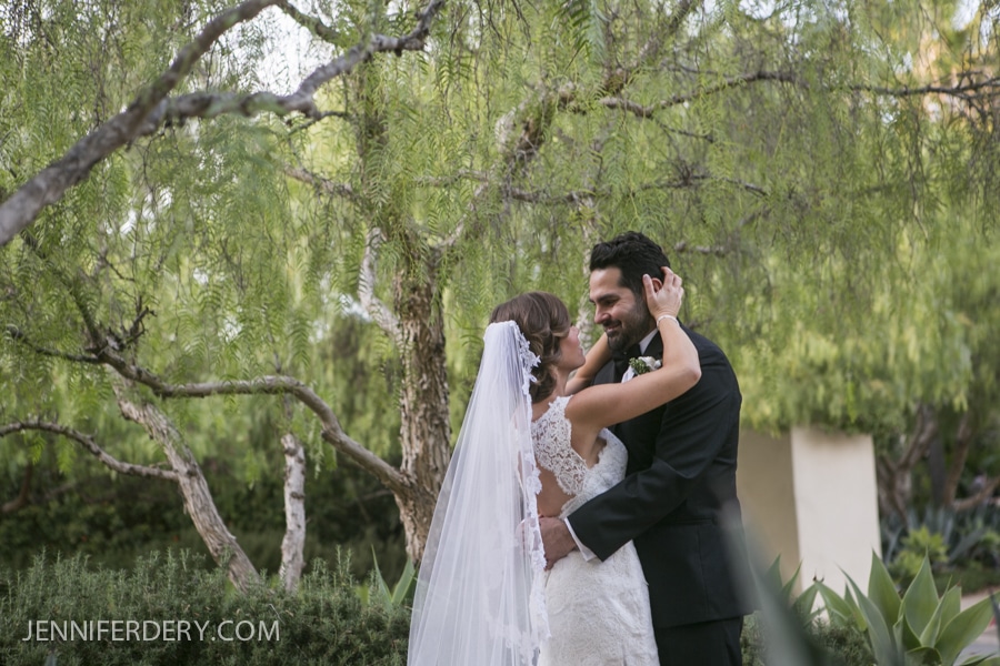 A bride and groom embrace and smile at each other outdoors during their Estancia La Jolla Wedding, surrounded by lush green trees and plants. The bride wears a veil and lace dress; the groom is in a black suit.