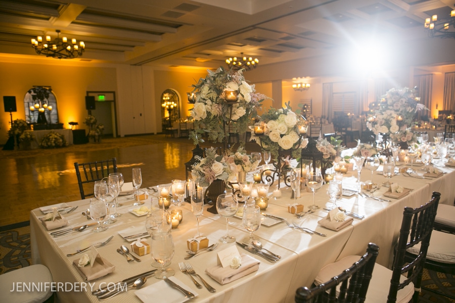 A long, elegant banquet table is set for an Estancia La Jolla Wedding, decorated with white flowers, candles, and neatly arranged place settings in a warmly lit, spacious ballroom with chandeliers and soft yellow lighting.