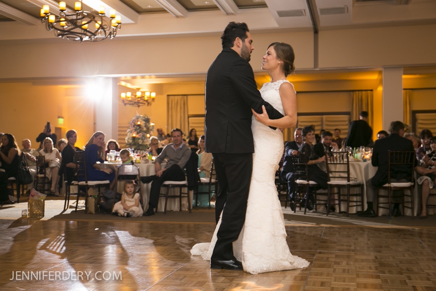 A bride and groom share their first dance in a warmly lit Estancia La Jolla Wedding reception hall, surrounded by smiling guests. A Christmas tree glows softly in the background.