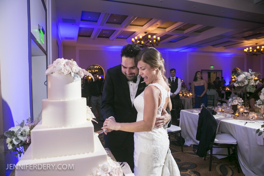A bride and groom, both smiling, cut a white tiered wedding cake together at their Estancia La Jolla Wedding reception in a softly lit banquet hall adorned with flowers.