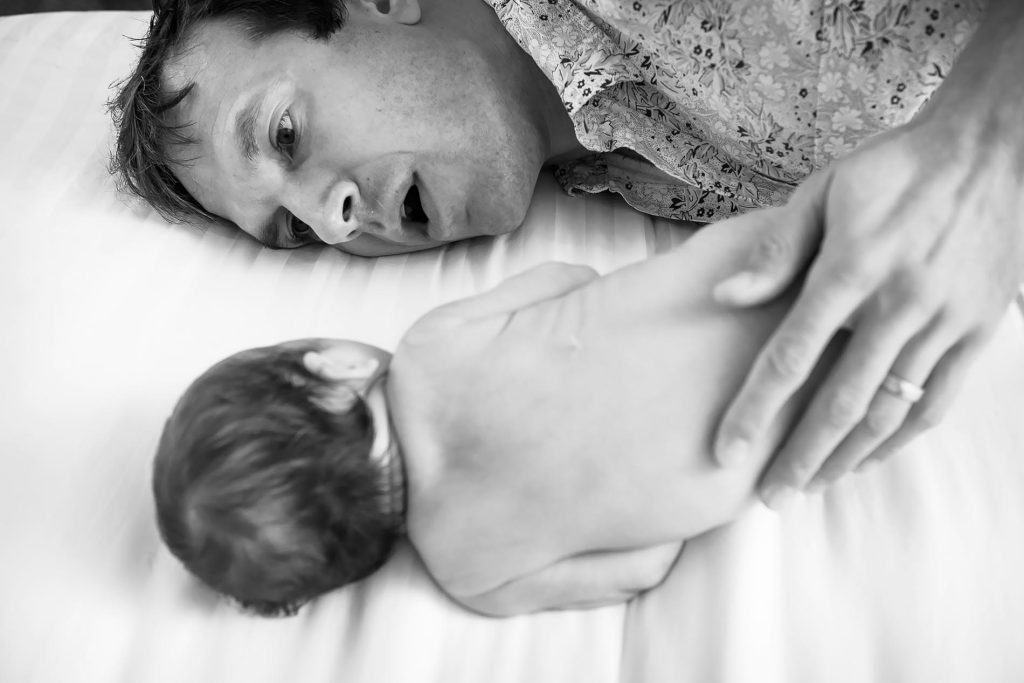 A black and white image shows a man lying on a bed, looking closely at an infant lying on its stomach. The man's expression is gentle and curious, while his hand gently supports the baby. The bed is covered in a white sheet.